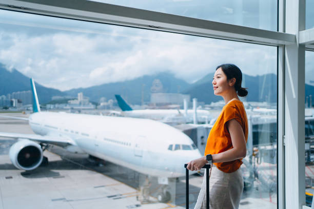 young asian woman carrying suitcase, looking through window at airport terminal while waiting for departure. ready for a trip. business travel. travel and vacation concept - travel photos et images de collection