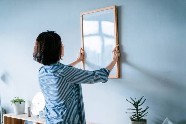young asian woman decorating and putting up a picture frame on the wall at home - home decoration stock pictures, royalty-free photos & images