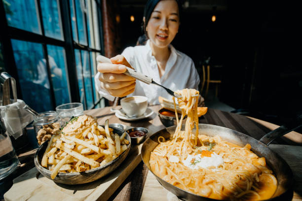 young asian woman sitting at a table by the window enjoying the warmth of sunlight and having meal joyfully in a restaurant - food stock pictures, royalty-free photos & images