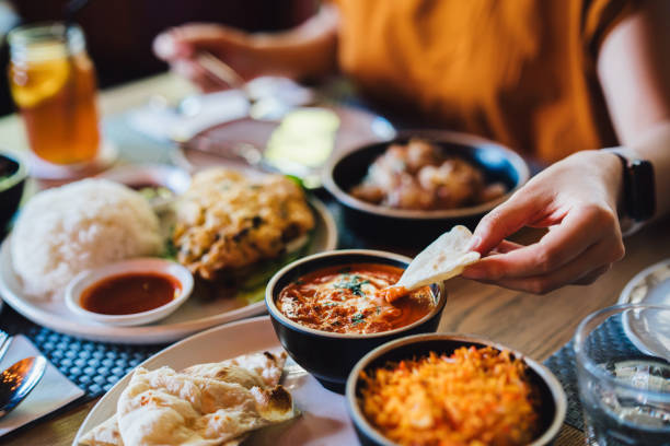 young asian woman tasting authentic classic indian food with assorted dishes freshly served on the table in restaurant. close-up of her hand dipping naan bread on the butter chicken. tourist exploring local cuisines when trav