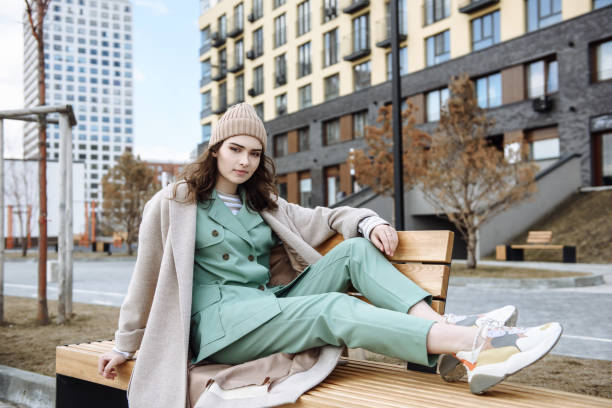 young beautiful teenage girl in a hat and coat sits on a bench and poses against the background of a city street - fashion stock pictures, royalty-free photos & images