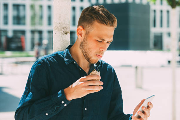 young businessman having lunch outside while looking at his cell phone - junk food stock pictures, royalty-free photos & images