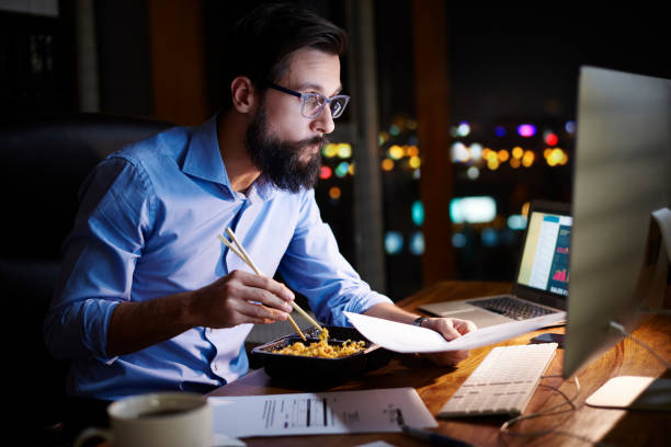 young businessman looking at computer and eating takeaway at office desk at night - junk food stock pictures, royalty-free photos & images