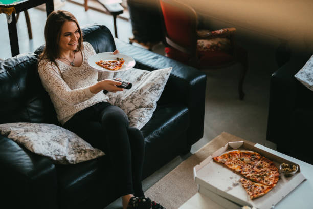 young casually dressed woman eating pizza in front of a tv - junk food stock pictures, royalty-free photos & images