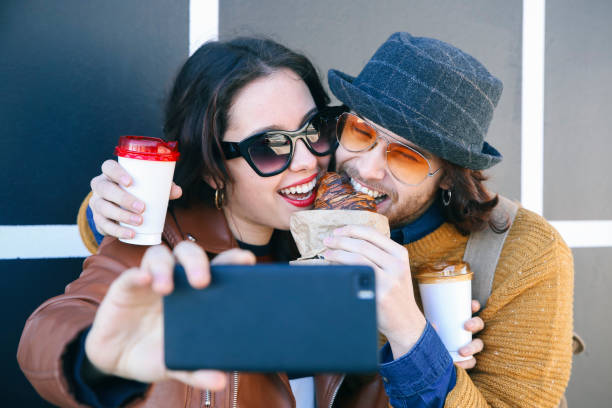 young couple with croissant and coffee to go taking selfie with smartphone - junk food stock pictures, royalty-free photos & images