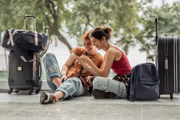 young friends woman using phone sitting on ground while waiting at airport - travel stock pictures, royalty-free photos & images