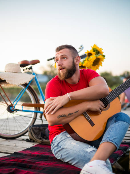 young hipster guy holding acoustic guitar while sitting on dock in nature. summer and travel concept. - concert stock pictures, royalty-free photos & images