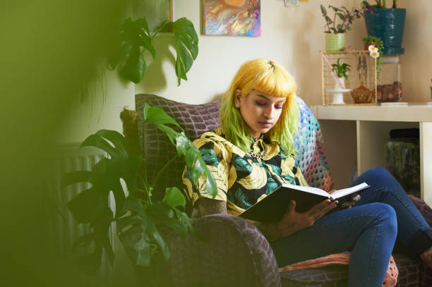 young hipster woman reading a book in her living room - home decoration photos et images de collection