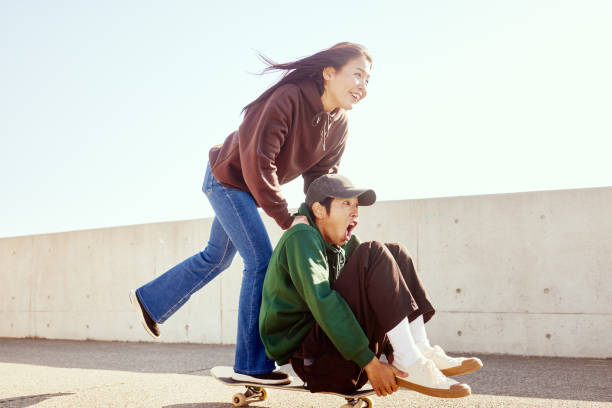 young man enjoys pushing his friend sitting on a skateboard from behind with his hand. - fashion stock pictures, royalty-free photos & images