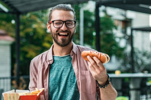 young man is eating hot dog on the street - junk food stock pictures, royalty-free photos & images