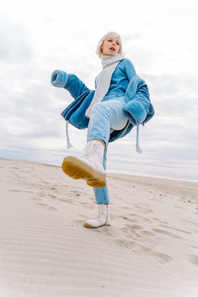 young modern blond woman in warm jeans outfit and a scarf posing on a sandy beach. sea background - fashion stock pictures, royalty-free photos & images