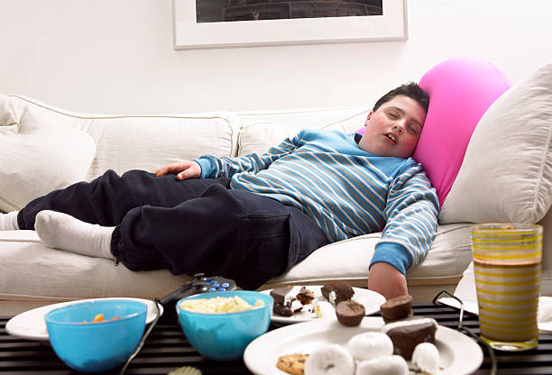 young, overweight boy sleeps on a sofa next to a table of crisps and biscuits - junk food stock pictures, royalty-free photos & images
