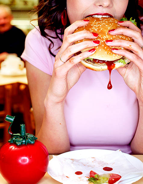 young woman biting into hamburger in diner - junk food stockfoto's en -beelden