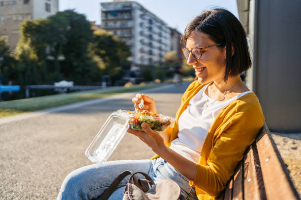 young woman eating a salad on a bench in the park - junk food stock pictures, royalty-free photos & images