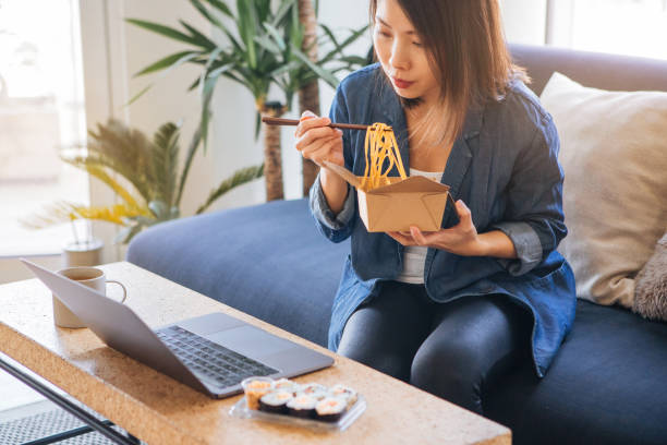 young woman eating takeaway food while using laptop - junk food stock pictures, royalty-free photos & images