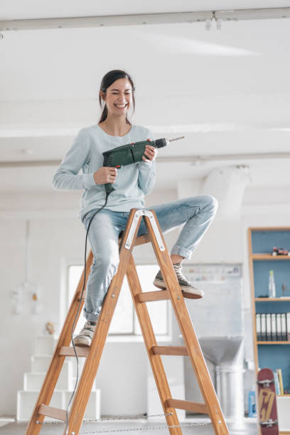 young woman in her new flat sitting on ladder, holding electric drill - home decoration stockfoto's en -beelden
