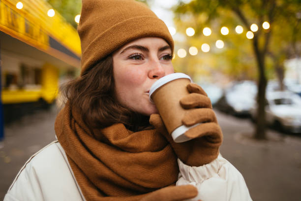 young woman in warm clothing drinking coffee on street during autumn - junk food stock pictures, royalty-free photos & images