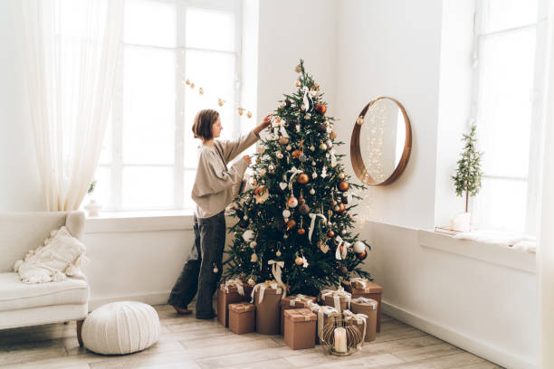 young woman is decorating christmas tree with many different decorations and festive garland in minimalist light interior. new year celebration concept - home decoration stockfoto's en -beelden