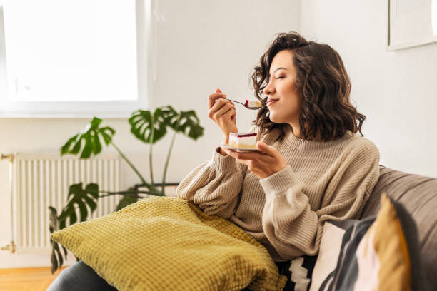 young woman is sitting in the living room alone enjoying taste of the cake - food stock pictures, royalty-free photos & images