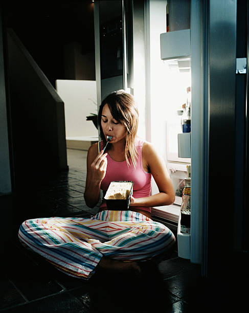 young woman sitting by open refrigerator, eating ice cream - junk food stock pictures, royalty-free photos & images