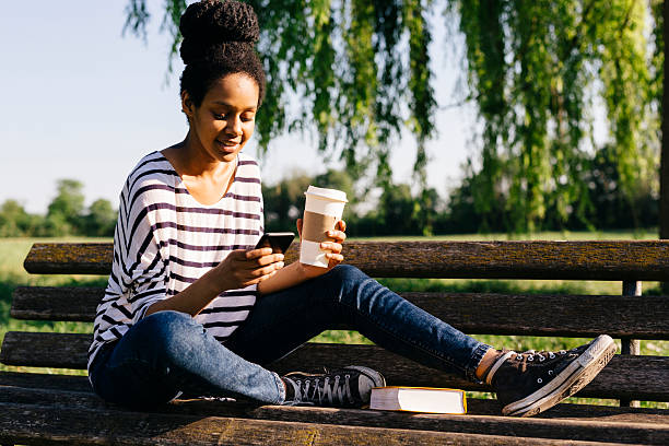 young woman sitting on park bench with a book and coffee to go looking at smartphone - junk food stock pictures, royalty-free photos & images