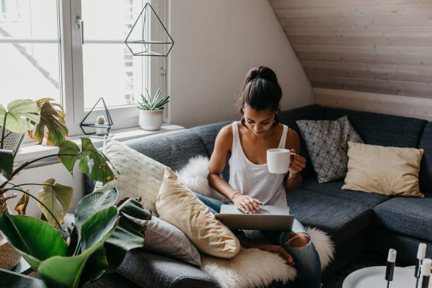young woman sitting on the couch with cup of coffee using laptop - home decoration fotografías e imágenes de stock