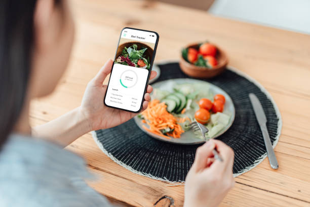 young woman using calorie counter app on smartphone while eating salad - food stock pictures, royalty-free photos & images