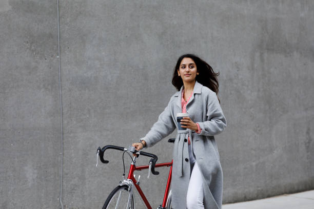 young woman walking with bicycle, with reusable coffee cup in hand - junk food stock pictures, royalty-free photos & images