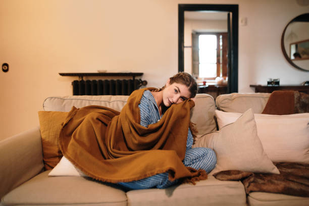young woman wearing pajamas and a blanket sitting on sofa - home decoration stock pictures, royalty-free photos & images