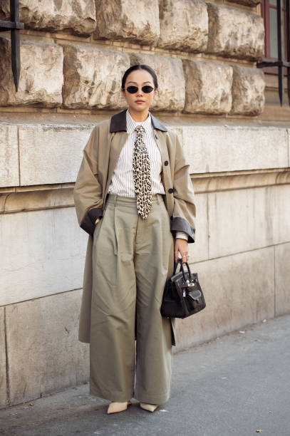 Yoyo Cao wears black sunglasses, a leopard printed tie, a white and beige striped shirt, a black leather Birkin Hermès bag, a beige long trench coat...