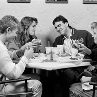 1980s GROUP OF FOUR TEENS SITTING AROUND TABLE AT RESTAURANT CHATTING & EATING HAMBURGERS
