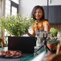 a beautiful asian woman florist is vlogging on her smartphone and laptop, creating an online tutorial on flower bouquet arrangement at home. this scene represents the concept of self-employment, e-learning, online classes, an