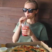 a beautiful stylish girl in sunglasses drinks a soft drink or cocktail through a straw, outdoors in a cafe. italian cuisine. sliced pizza in paper packaging on a wooden table. delicious lunch, snack, fast food. - junk food st