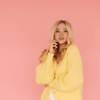 a beautiful young blonde woman with curly hair in a yellow knitted cardigan is chatting on the phone, communicating using a mobile phone while standing on a pink isolated background. place for text - fashion stock pictures, r