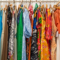 a colorful selection of vibrant dresses and garments hanging on a clothing rack, featuring an array of bold patterns and bright hues, from polka dots and floral prints to abstract designs from a second-hand vintage shop. spai