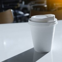 a disposable glass or mug with hot coffee or tea to take away on a white table in a modern stylish youth cafe, cafeteria or restaurant. food and beverage establishment. fast food restaurant. the shadow of the mug. breakfast a