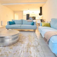 a living room set, light blue furniture and blue cushions, a round silver mousetrap table, a tv and a picture on the side, in the background you can see the kitchen and two lit lamps, an exit sign from the apartment. - home d