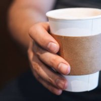 a person holding a takeaway coffee cup in hand with a cardboard sleeve, captured in a cozy indoor setting during the morning - junk food stock pictures, royalty-free photos & images