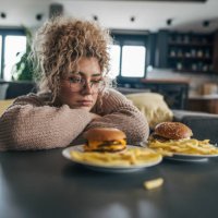 a plus-sized woman with curly hair sitting on the floor, gazing at the two plates of hamburgers and french fries on the table - junk food stock pictures, royalty-free photos & images