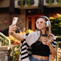 a smiling woman in sunglasses and wireless headphones takes selfies using a mobile phone and listens to music while walking along an autumn street in the countryside outdoors - fashion stock pictures, royalty-free photos & im