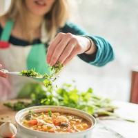 a woman wearing an apron, sitting at a table, sprinkling herbs into a bowl of vegetable stew. - food stock pictures, royalty-free photos & images