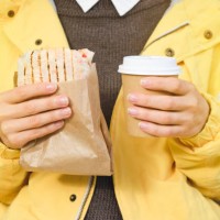 a young woman or girl holding a cup of coffee, drinking it, eating a delicious shawarma doner kebab, sitting on a bench in a public park. fast food and snack concept. - junk food stock pictures, royalty-free photos & images