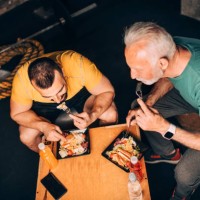active senior sportsman eating a healthy protein salad meal in a take-out box with his personal trainer in a gym area of a gym - junk food stock pictures, royalty-free photos & images