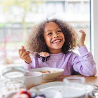 adorable black girl with curly hair smiling while eating breakfast in a bright kitchen - food stock pictures, royalty-free photos & images