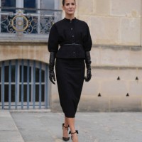 Alex Riviere wears black puffy short sleeve tailored Dior blazer jacket, black leather belt, black Dior midi skirt, shiny black leather gloves, black...