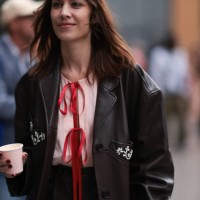 Alexa Chung is seen wearing a knee-length black leather jacket with silver gemstone details, underneath a rose top with red ribbons and a black...