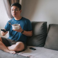 an asian chinese mid adult man having his take out food while pressing tv remote control watching tv on sofa - junk food stock pictures, royalty-free photos & images