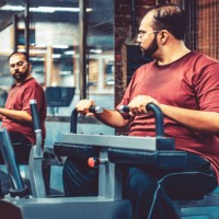 an overweight man at a gym using exercising machine while looking at the mirror - junk food stock pictures, royalty-free photos & images