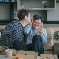 asian chinese man kissing his down syndrome son enjoying pizza fast food in living room sitting on sofa during weekend - junk food stock pictures, royalty-free photos & images