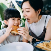 asian family having fun while enjoying lunch in an outdoor restaurant, mother and daughter are sharing and having a big bite eating a burger. family eating out lifestyle - junk food stock pictures, royalty-free photos & image